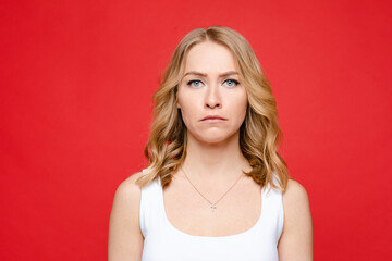 Young beautiful caucasian woman with medium fair wavy hair and nude makeup in white t-shirt looks with disbelief, picture isolated on red background