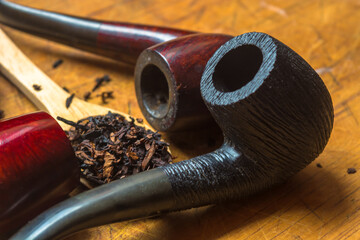 Wooden spoon with a heap of tobacco blend in the middle of three classic red, black and brown smoking pipes on a wooden yellow table.