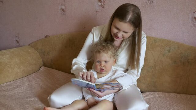 Beautiful attractive mother with a cute baby sit at home on the couch and have fun looking at the picture book.