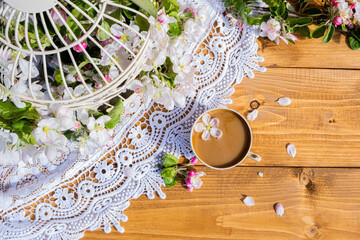 A cup of coffee with a fallen apple tree flower on a wooden table with a lace napkin. The metal cage is decorated with branches of a blossoming apple tree. Healthy breakfast concept. Interior concept.