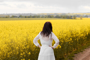 Fototapeta premium Woman in a white dress standing back in a yellow rapeseed field. Spring concept. 