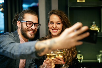 Smiling couple sitting in restaurant toasting with white wine and taking selfie.