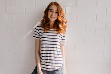 Wonderful lady in striped t-shirt looking to camera with interest. Shy positive girl standing on white background.