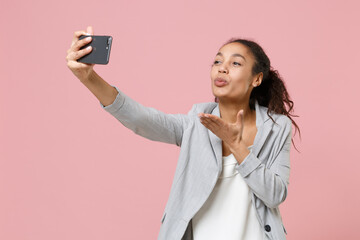 Pretty african american business woman in grey suit, white shirt isolated on pink background. Achievement career wealth business concept. Doing selfie shot on mobile phone, blowing sending air kiss.