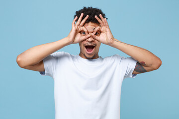 Excited young african american guy in casual white t-shirt posing isolated on blue background. People lifestyle concept. Mock up copy space. Holding hands near eyes, imitating glasses or binoculars.