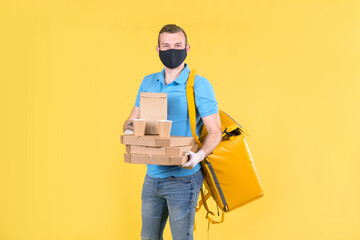 Young food delivery guy in protective mask and gloves is holding an order from restaurant for customer, dressed in blue Polo shirt and carrying yellow shopping bag on his shoulders. Safe food delivery