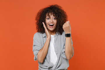 Fototapeta na wymiar Excited young african american woman in gray casual clothes isolated on orange background studio portrait. People lifestyle concept. Mock up copy space. Wearing smart watch on hand put hand on cheek.