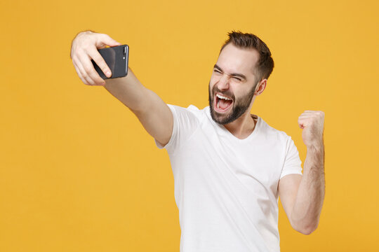 Joyful young bearded man guy in white casual t-shirt posing isolated on yellow wall background. People lifestyle concept. Mock up copy space. Doing selfie shot on mobile phone, doing winner gesture.