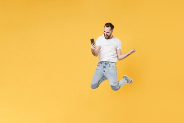 Fototapeta na wymiar Happy young bearded man guy in white casual t-shirt posing isolated on yellow background. People lifestyle concept. Mock up copy space. Jumping doing selfie shot on mobile phone doing winner gesture.