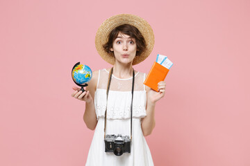 Amazed young tourist girl in summer dress hat with photo camera isolated on pink background. Traveling abroad to travel weekend getaway. Air flight journey concept. Hold passport tickets world globe.