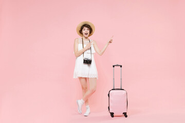 Excited tourist girl in summer dress hat with photo camera suitcase isolated on pink background. Female traveling abroad to travel weekend getaway. Air flight journey concept. Point index fingers up.
