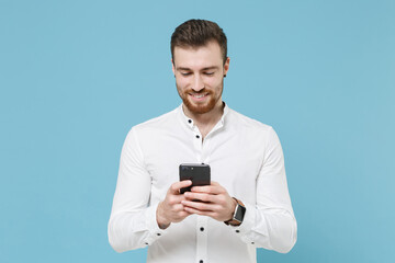 Smiling young bearded man guy 20s in white classic shirt isolated on pastel blue wall background studio portrait. People lifestyle concept. Mock up copy space. Using mobile phone, typing sms message.