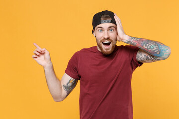 Amazed young bearded tattooed man guy in casual t-shirt black cap posing isolated on yellow background. People lifestyle concept. Mock up copy space. Pointing index finger aside, put hand on head.