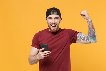 Cheerful young bearded tattooed man guy in casual t-shirt black cap posing isolated on yellow wall...