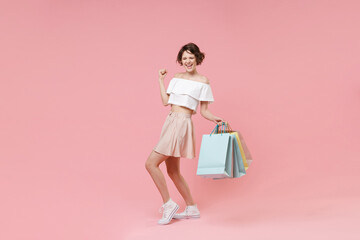 Joyful young woman girl in summer clothes hold package bag with purchases isolated on pastel pink background in studio. Shopping discount sale concept. Mock up copy space. Clenching fists like winner.