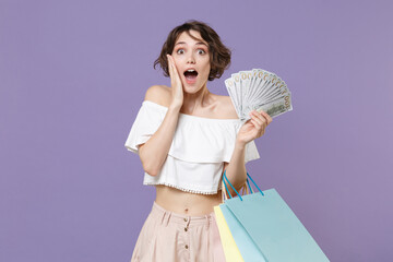Shocked young woman girl in summer clothes isolated on violet wall background. Shopping discount sale concept. Hold package bag with purchases fan of cash money in dollar banknotes put hand on cheek.