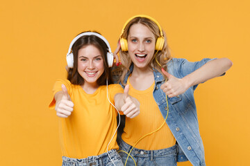 Two cheerful young women girls friends in casual t-shirts denim clothes isolated on yellow background. People lifestyle concept. Mock up copy space. Listen music with headphones, showing thumbs up.