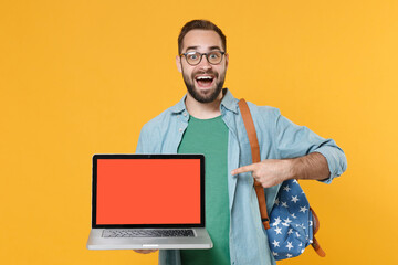 Excited young man student in glasses with backpack isolated on yellow background. Education in high school university college concept. Point index finger on laptop pc computer with blank empty screen.