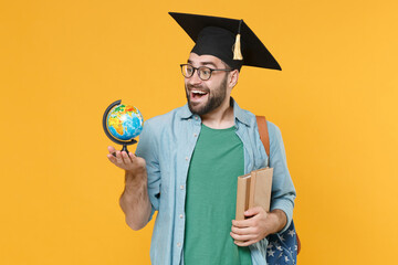 Excited young man student in graduation cap glasses with backpack hold books world globe isolated...