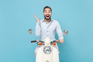 Surprised young bearded man guy in casual light shirt driving moped isolated on blue wall background. Driving motorbike transportation concept. Mock up copy space. Keeping mouth open, spreading hands.