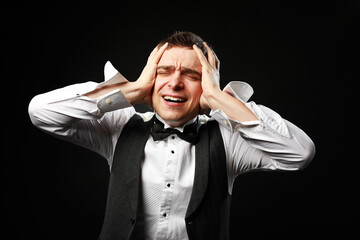 Portrait of young man in black bow tie isolated on black background suffering from severe headache, pressing fingers to temples, closing eyes to relieve pain with helpless face expression. 