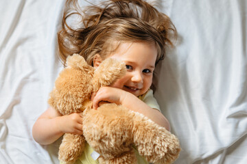 Smiling toddler girl holding teddy bear and lying in bed on white sheets at home. Happy child. Top view.