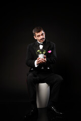 Young handsome man in tuxedo and bow tie siting on white bar chair and holding decor moss isolated on black background