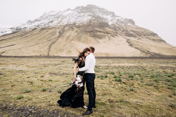 Wedding couple on a background of snowy mountains. The bride in a black dress and groom are hugging in a field of moss and yellow grass. Destination Iceland wedding. 
