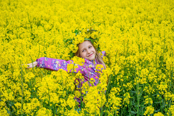 Fototapeta premium Happy smiling girl with closed eyes is standing in the field of yellow flowers. Rural landscape of blooming rapeseed.