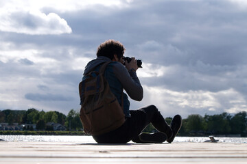 happy young man carrying brown backpack taking photos sitting in a quiet place on the edge of a dock on a large lake with a view of a large sky with gray clouds - Powered by Adobe