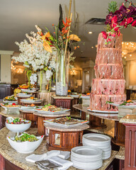 Large variety of food on a table at a formal event with flowers and a tier of prosciutto.