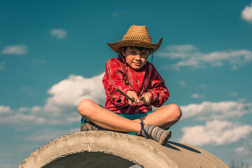 happy funny boy, a child in a straw hat and a stick, sits on a concrete ring against the sky and clouds. nature is a clear and sunny day