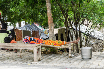 Adult wearing colorful clothing, sleeping on a cot in a poverty region of Agra, India, with tents in the background.