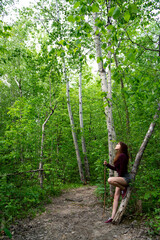 Naklejka na ściany i meble Happy redhead woman wearing cherry shirt and shoes and gray shorts looking up resting on a trail on a tree trunk holding a branch in the middle of the deep green leafy forest