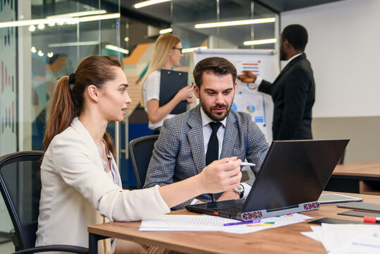 Joined work of two confident business people in formal suit working together with laptop at office with their coworkers at background.
