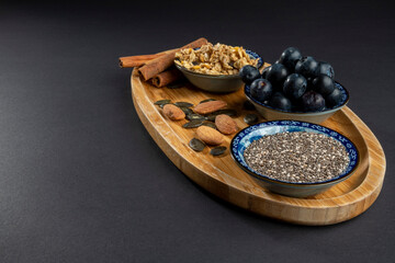 Healthy/natural food concept: On wooden board, some cinnamon sticks, almonds and sunflower seeds. In blue bowls: crunchy granola, blueberries and chia seeds. Black background with copy space