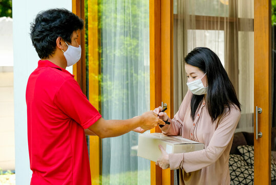 Beautiful Woman With Mask Get Delivery Package Box From Carrier Man With Red Shirt And Mask Then Sign At Home. Concept Of New Normal Lifestyle To Prevent Virus Infection During Covid Pandemic In City.