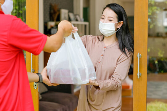 Beautiful woman with mask get delivery pizza or food from carrier man with red shirt and mask at home. Concept of new normal lifestyle to prevent virus infection during covid pandemic in city.