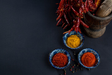 Obraz na płótnie Canvas Top view on three blue bowls with whole, granular and powdered red pepper and yellow mixed ginger, curry, turmeric powder and peppercorns. Still life on black background