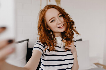 Amazing girl in striped t-shirt making selfie at home. Gorgeous ginger young woman posing in white headphones.