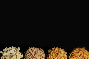 Different types of dry pasta on the plate and in bowls on black background. Space for text, top view