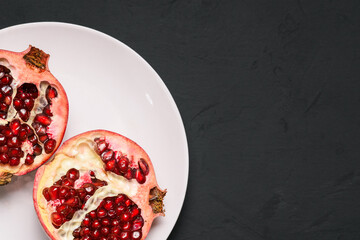 Cut into two parts ripe pomegranate on a white plate. Red garnet with grains on a black background with texture. Top view, copy space
