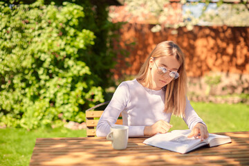 Fototapeta na wymiar A cute young teenage girl is reading a book while sitting at a wooden table with a Cup of coffee, in the back yard.