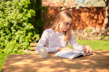 Fototapeta na wymiar A cute young teenage girl is reading a book while sitting at a wooden table with a Cup of coffee, in the back yard.