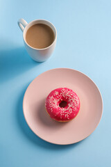 Pink donut with sprinkles on plate near to cup of coffee, sweet glazed dessert food and hot drink on blue minimal background, angle view