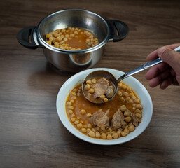 Traditional Turkish food chickpea in  saucepan with ladle and dish on brown table. Hand serving chickpea soup.