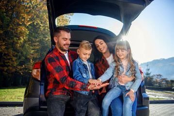 Adorable parents with their lovely kids sitting in the trunk of their family car, laughing and giving five to each other. Enjoying happy family time concept.