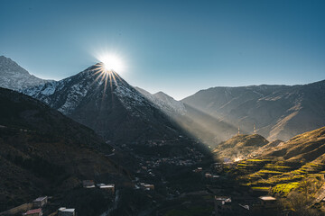 A view of sunset sunrise with sunstar in rural mountain village Imlil in High Atlas mountains Morocco in Africa. Snow covered peaks in background with lightrays and blue sky.