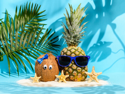 Cute fruit couple coconut with eyes and pineapple in sunglasses on beach sand among sea stars and palm leaves on blue background. Strong tropical shadows. Hawaiian vacation, summer concept