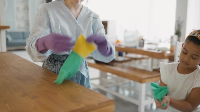 Young, caring mother of housewife wearing glasses in kitchen with her african american daughter wearing rubber gloves to protect her hands from detergents. Preparing for cleaning.